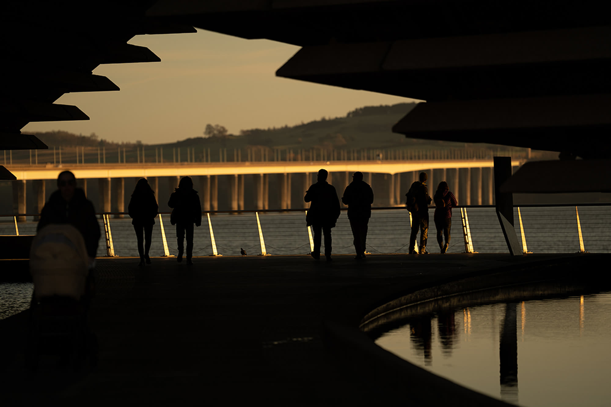 Silhouettes on the Dundee waterfront at sunset, the Tay Rail Bridge beyond, framed by the roof of V&A Dundee.